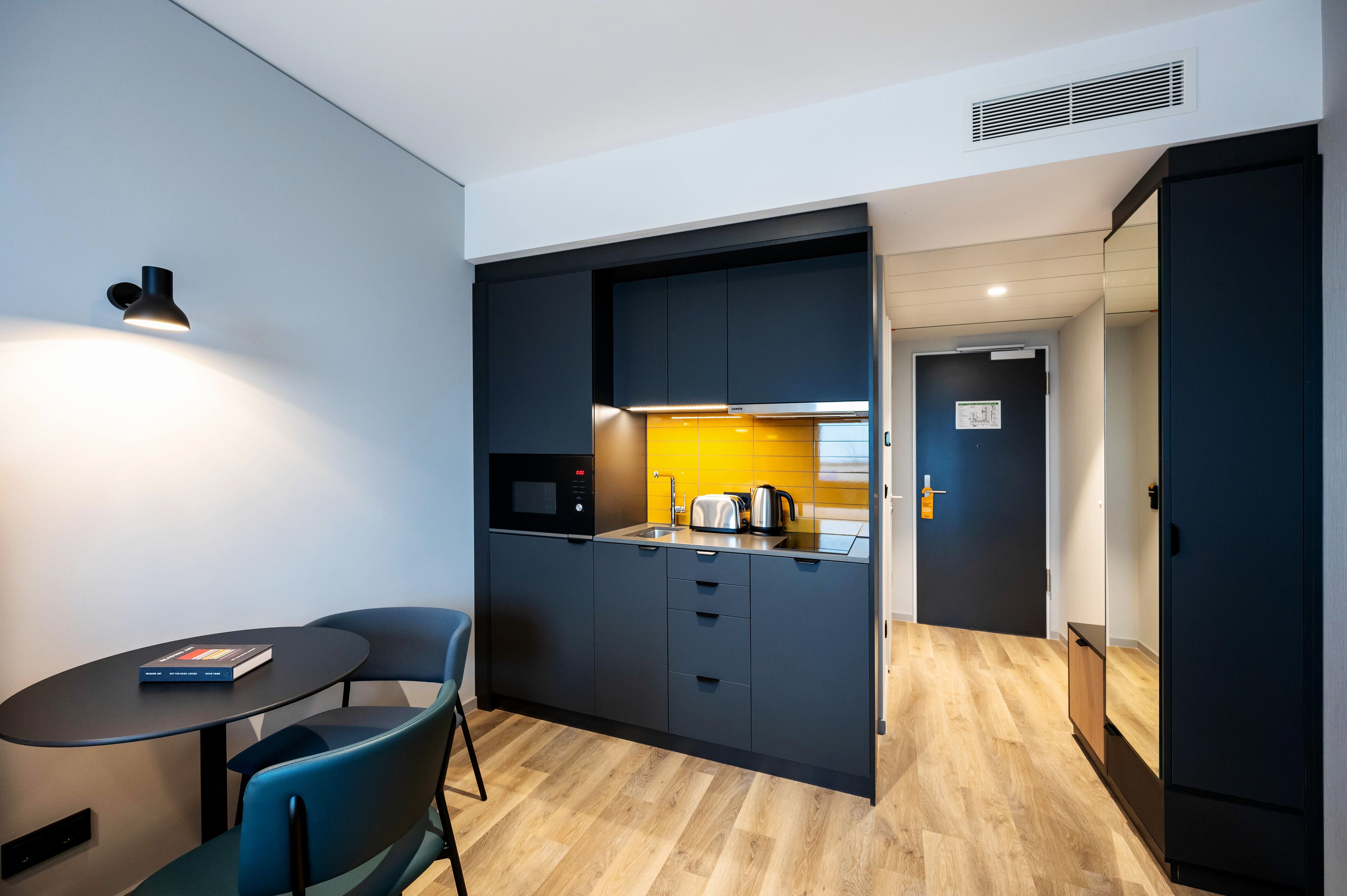 A compact kitchen with a microwave, toaster, and kettle, a small dining area with a table and chairs, and a dark-colored wardrobe.