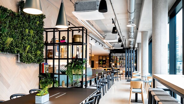 Um café moderno com um balcão comprido, cadeiras pretas e uma parede verde com plantas.