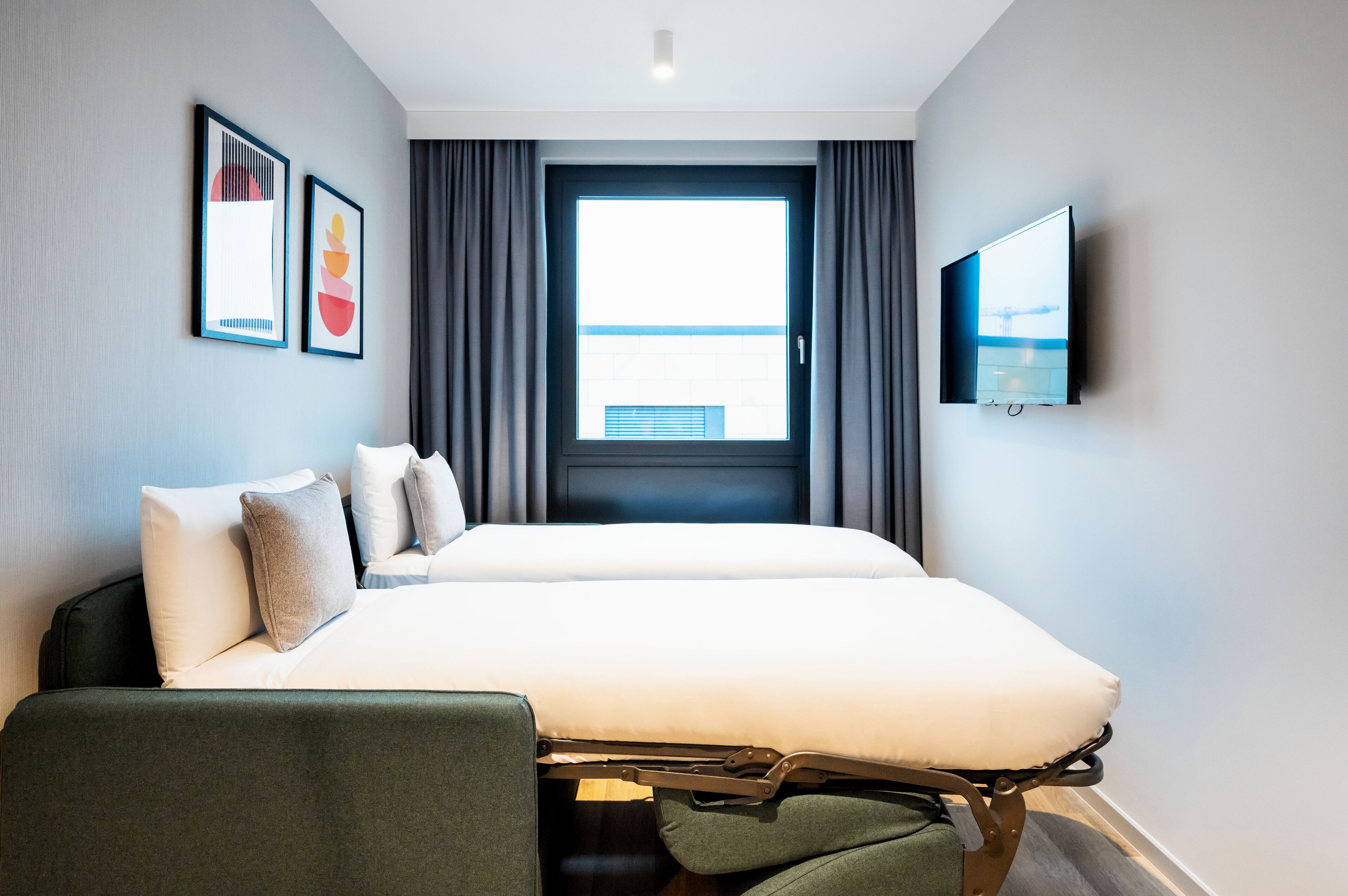 Une chambre d’hôtel avec un lit, une télévision, une fenêtre avec des rideaux et deux tableaux encadrés au mur.