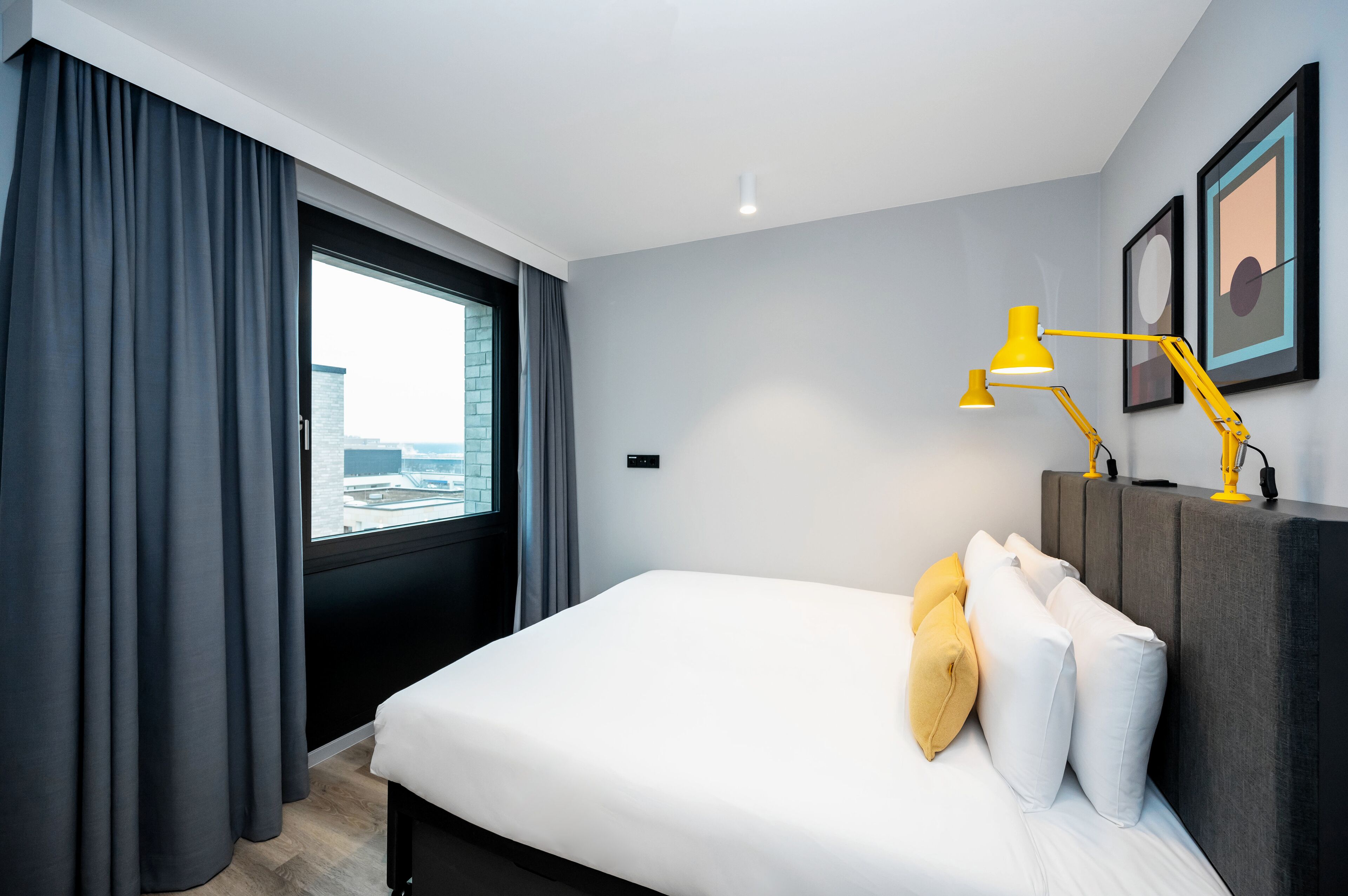 A modern hotel room with a large bed, a desk with a yellow lamp, grey curtains, and framed artwork on the wall.