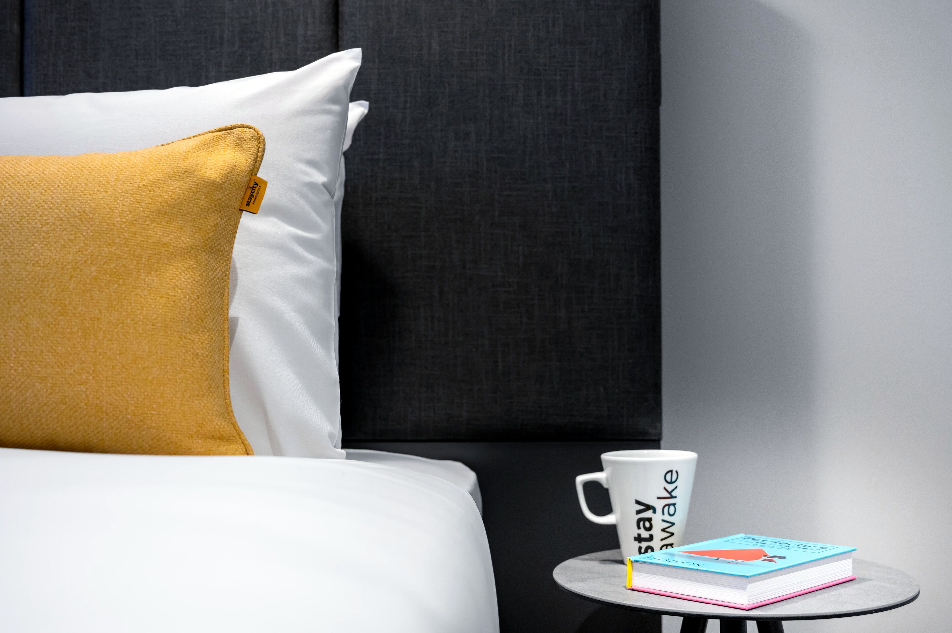 A neatly made bed with a white and mustard yellow pillow, a white mug with text, and a book on a small round table.