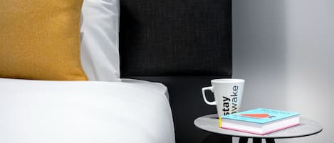 A neatly made bed with a white and mustard yellow pillow, a white mug with text, and a book on a small round table.