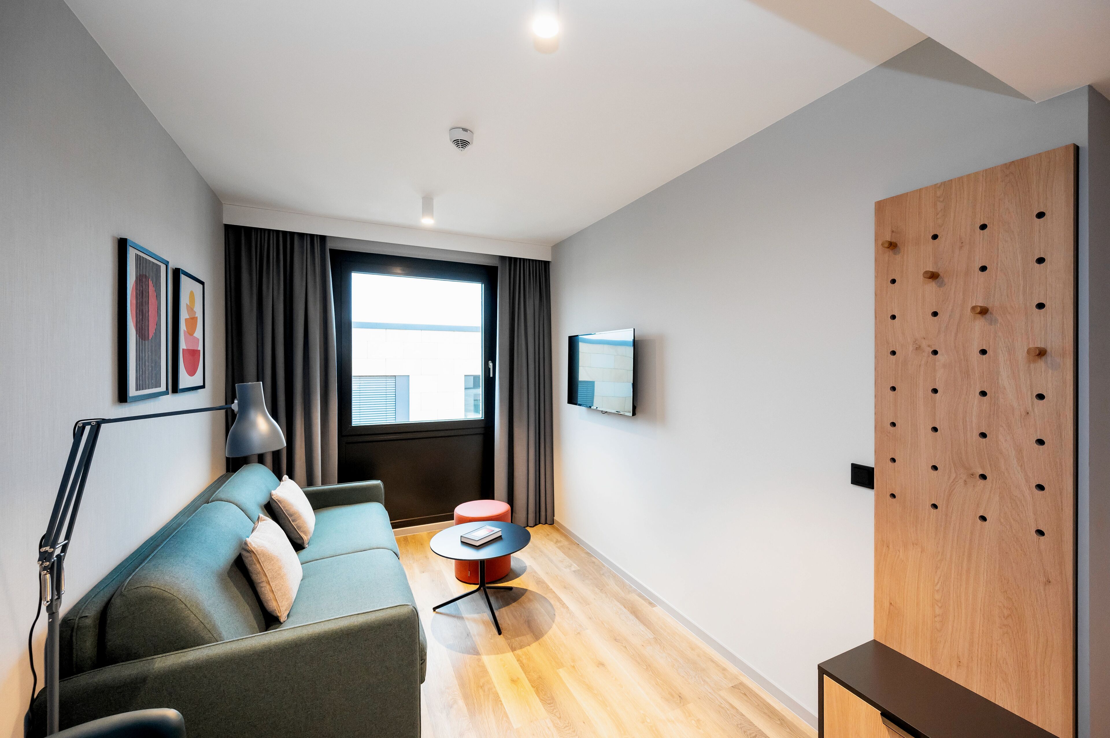 Una habitación de hotel moderna con un sofá, una pequeña mesa redonda, una lámpara, un televisor montado en la pared y un panel de madera con agujeros.