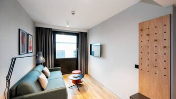 A modern hotel room with a sofa, a small round table, a lamp, a TV mounted on the wall, and a wooden panel with holes.
