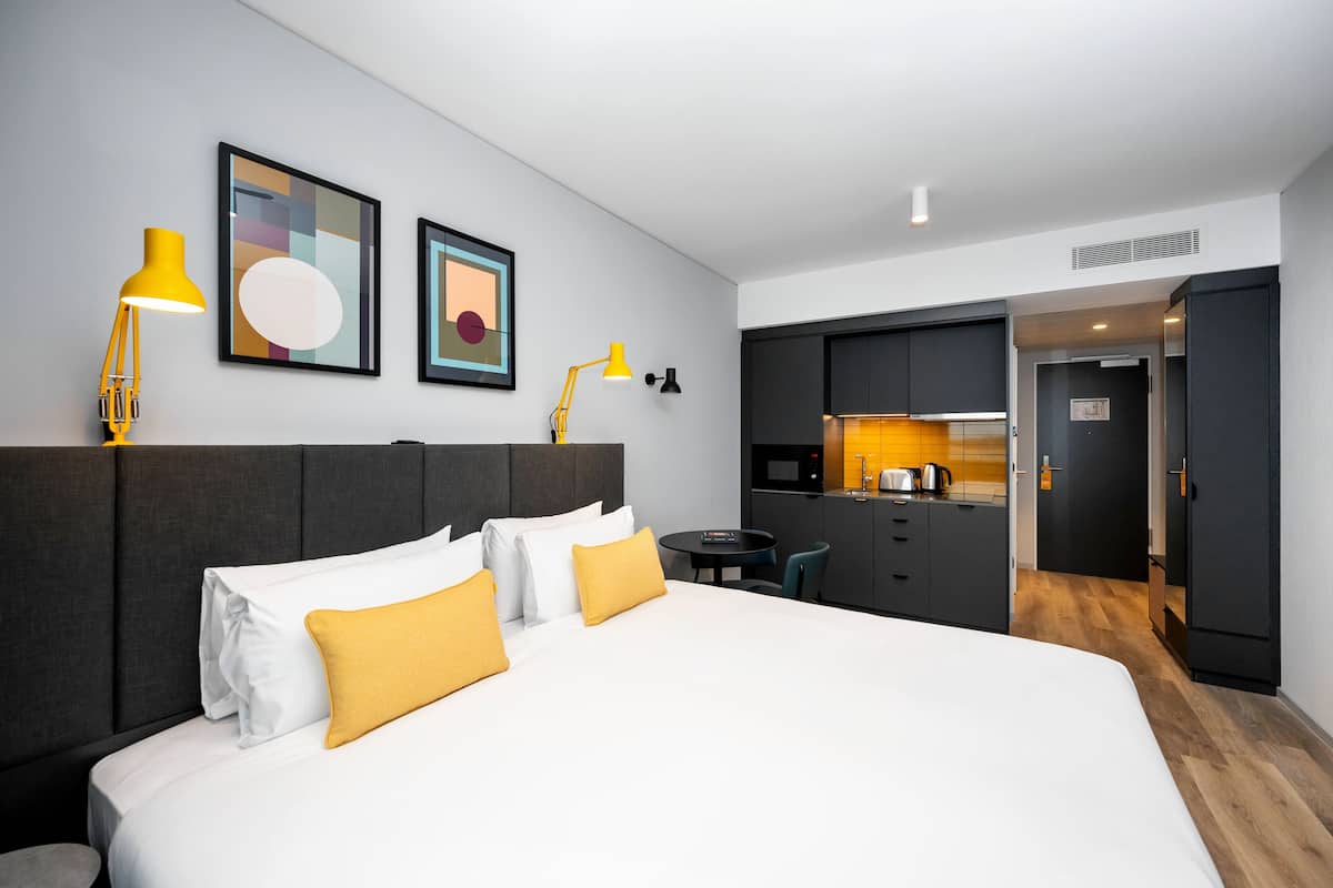 A modern hotel room with a large bed, a small desk, and a kitchenette.