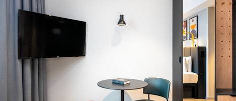 Habitación de hotel moderna con televisor de pantalla plana, una mesita redonda, dos sillas, un reposapiés rojo y un espejo que refleja un dormitorio con cama y obras de arte.