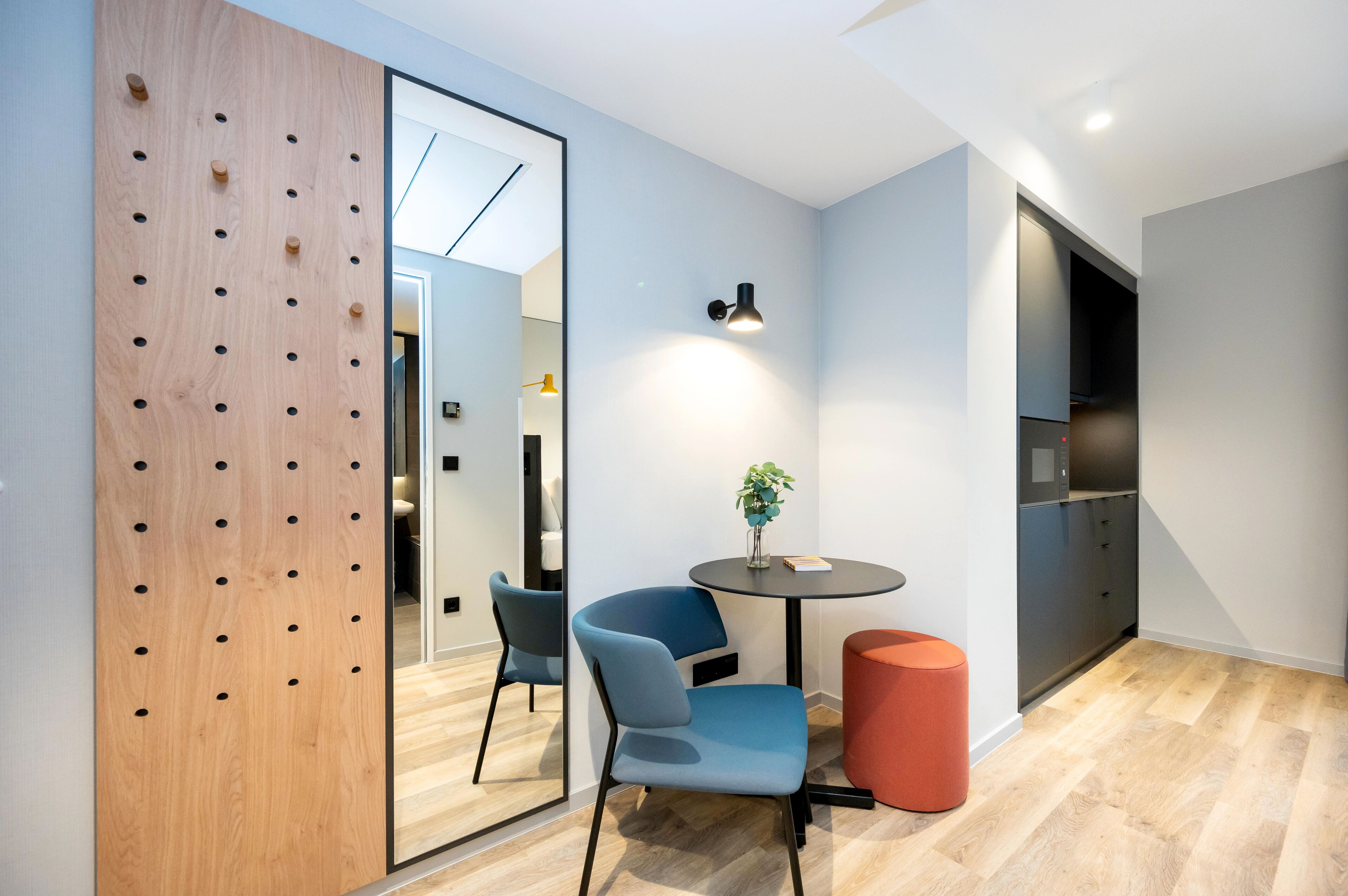 Una habitación moderna con un separador de madera, dos sillas, una mesita con una planta y un reposapiés rojo.