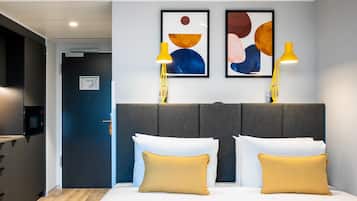A modern hotel room with a grey headboard, two yellow bedside lamps, and abstract artwork on the wall.