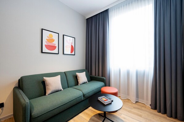 A modern living room with a green sofa, a round orange ottoman, and a black coffee table. There are two framed artworks on the wall and a large window with dark curtains.