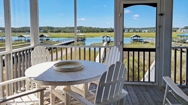 A covered outdoor area with a table and chairs, overlooking a water body and distant houses. - Creek Front Beach House, 3 Screened Porches, Private Dock, Easy Walk to Beach (Pawleys Island)