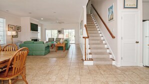 A spacious living area with a dining table, green sofas, and a staircase leading to another level. - Creek Front Beach House, 3 Screened Porches, Private Dock, Easy Walk to Beach (Pawleys Island)