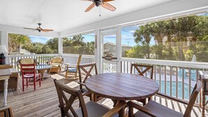 Un espace extérieur abrité, doté d’une terrasse en bois, d’une table ronde et de chaises, et offrant une vue sur une piscine et des palmiers.