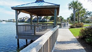 A wooden pier leading to a gazebo with a metal roof, overlooking a calm body of water and a landscaped area with palm trees.