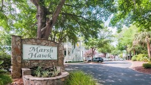 A sign for 'Marsh Hawk' with a stone base, surrounded by greenery and a residential street.
