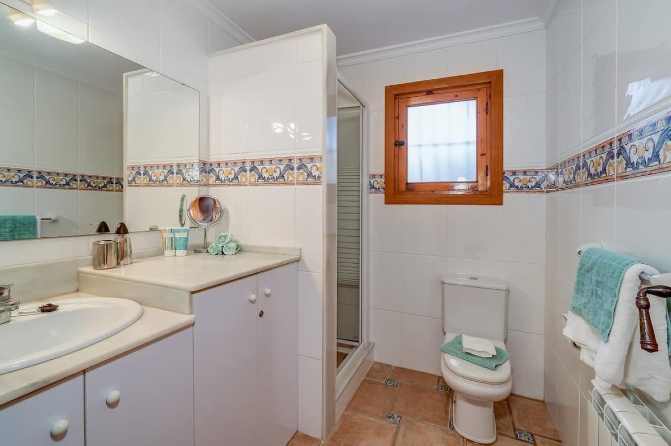 Un baño con lavamanos blanco, espejo y retrete. Hay una ventana sobre el retrete y un perchero con toallas.