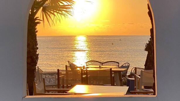 Vista do pôr do sol através de uma janela em arco, com vista para uma praia e uma mesa com cadeiras.