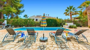 Área da piscina com espreguiçadeiras e um guarda-sol, cercada por vegetação e um edifício ao fundo.