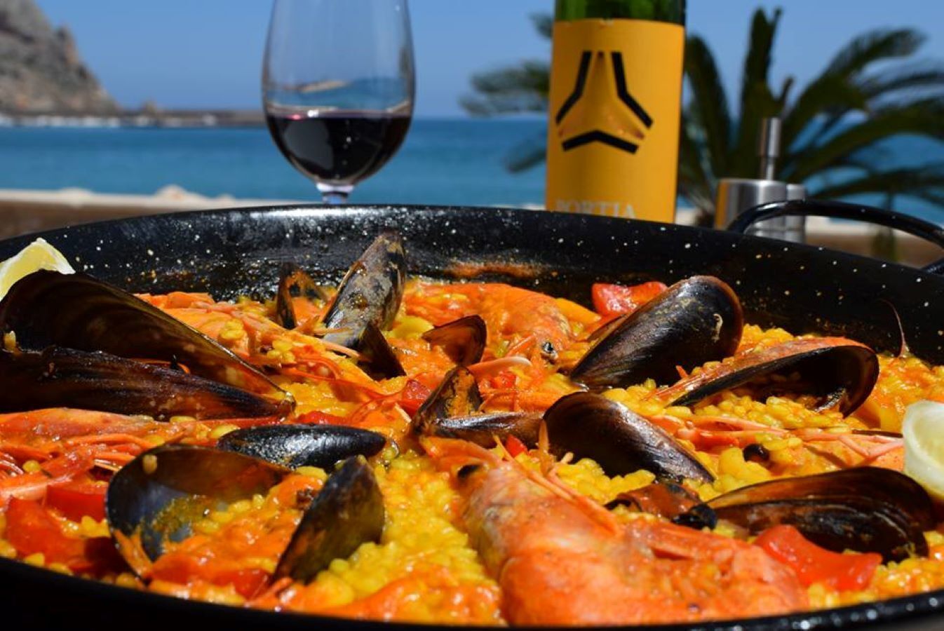 Paella mit Meeresfrüchten, darunter Miesmuscheln und Garnelen, ein Glas Rotwein und eine gelbe Flasche mit schwarzem Etikett.