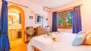 Quarto de hotel com cama, escrivaninha, cadeira, janela com cortinas e banheiro visível por uma arcada.