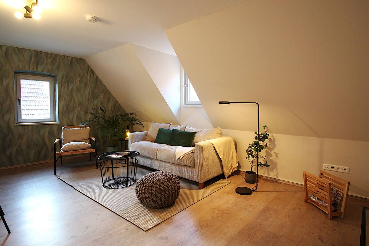A cozy loft bedroom with a sloped ceiling, a large window, a sofa, a coffee table, a floor lamp, and a potted plant.