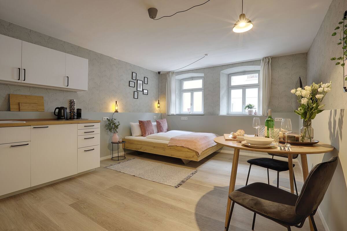 A modern, compact living space with a kitchenette, a bed, a dining area, and a large window providing natural light.