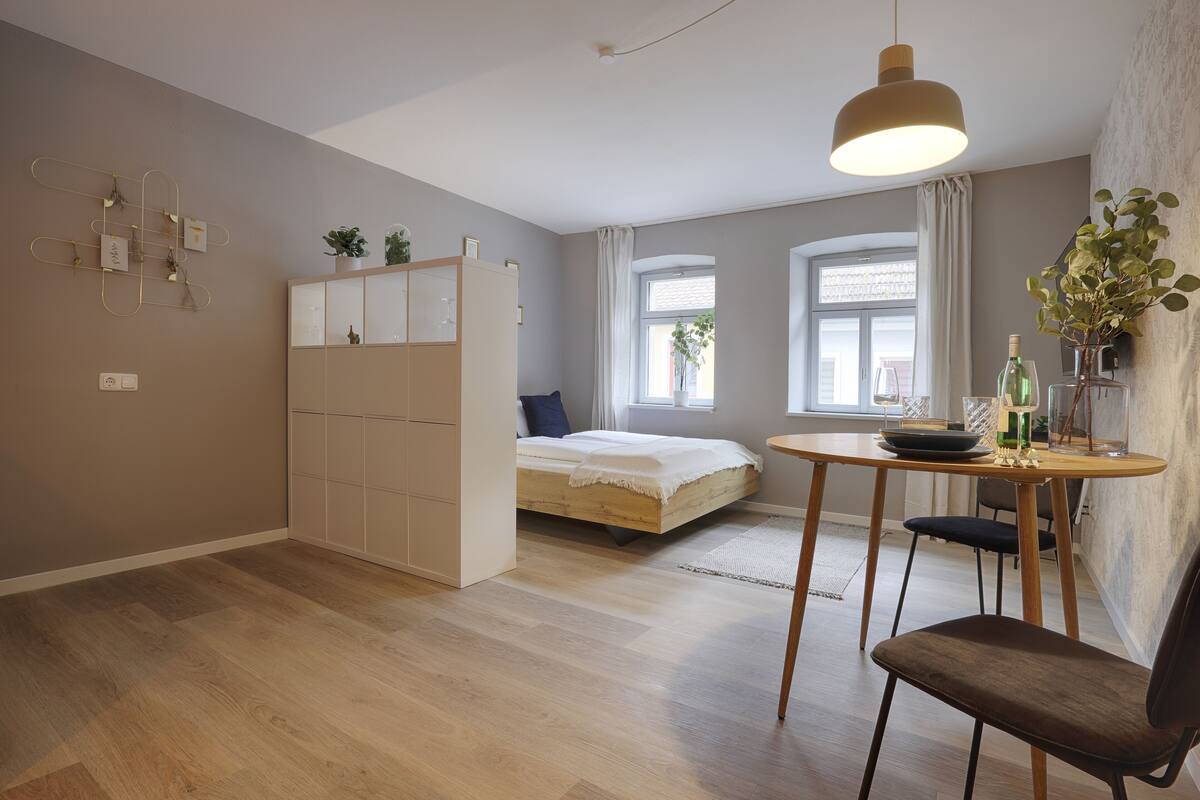 A modern bedroom with a wooden bed, a built-in wardrobe, a dining table with chairs, and a pendant light.