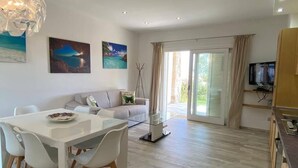 A modern living room with a white sofa, dining table, and chairs. There are paintings on the wall, a television, and a large window with curtains. - R House 2 independent semi-detached house (Budoni)