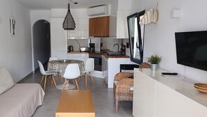 A modern living room with a sofa, dining table, and chairs. There is a kitchen area with cabinets, a stove, and a sink. A TV is mounted on the wall, and there is a window with curtains.