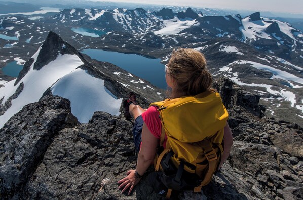 A person sitting on a rocky outcrop overlooking a mountainous landscape with snow-capped peaks and a lake. - Filefjellstuene (Vang)