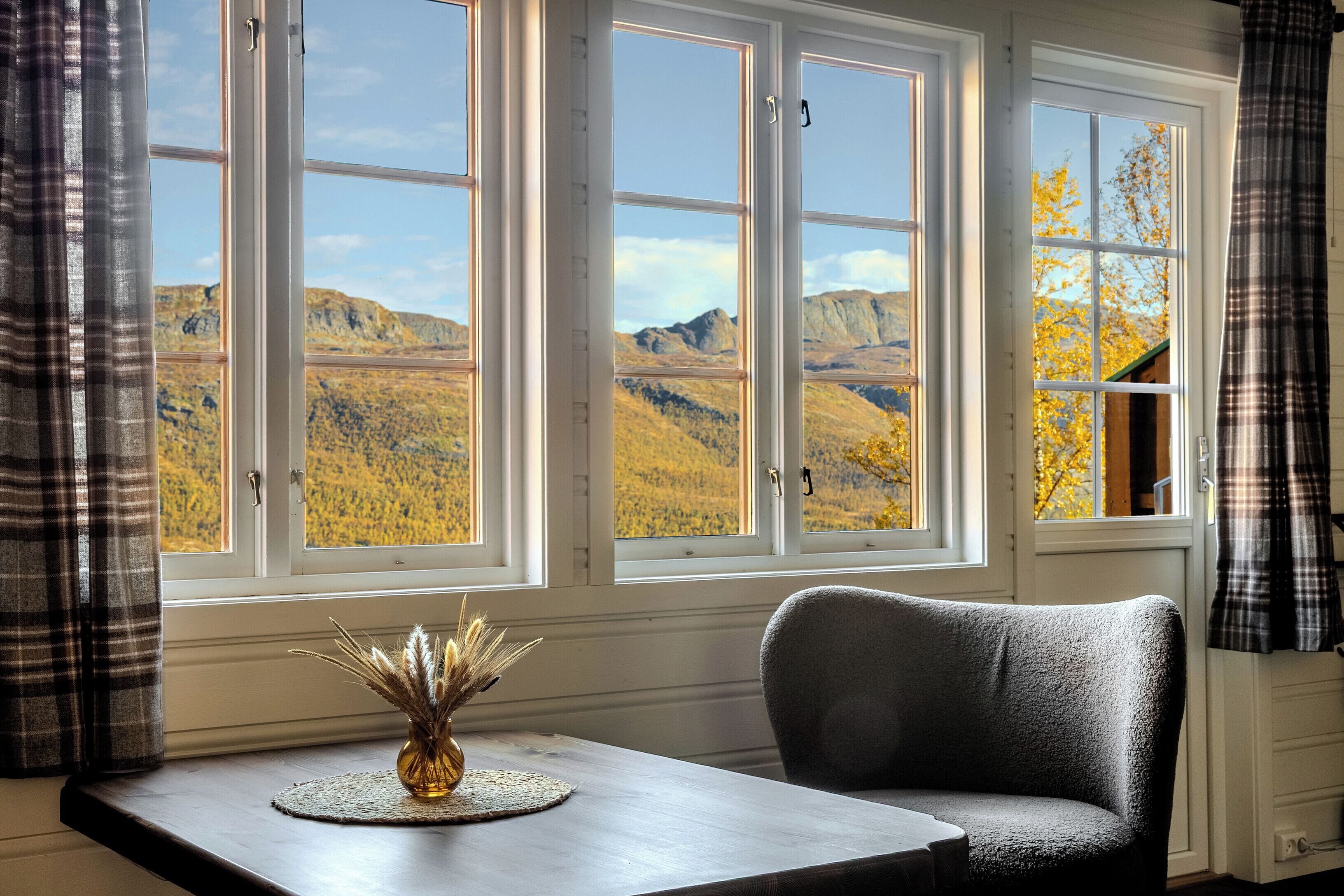 Habitación con vistas a montañas a través de amplios ventanales, una mesa con un florero y una silla gris.