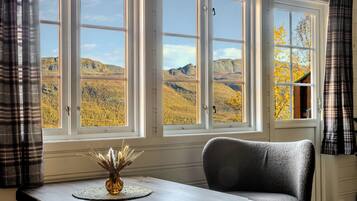 Habitación con vistas a montañas a través de amplios ventanales, una mesa con un florero y una silla gris.