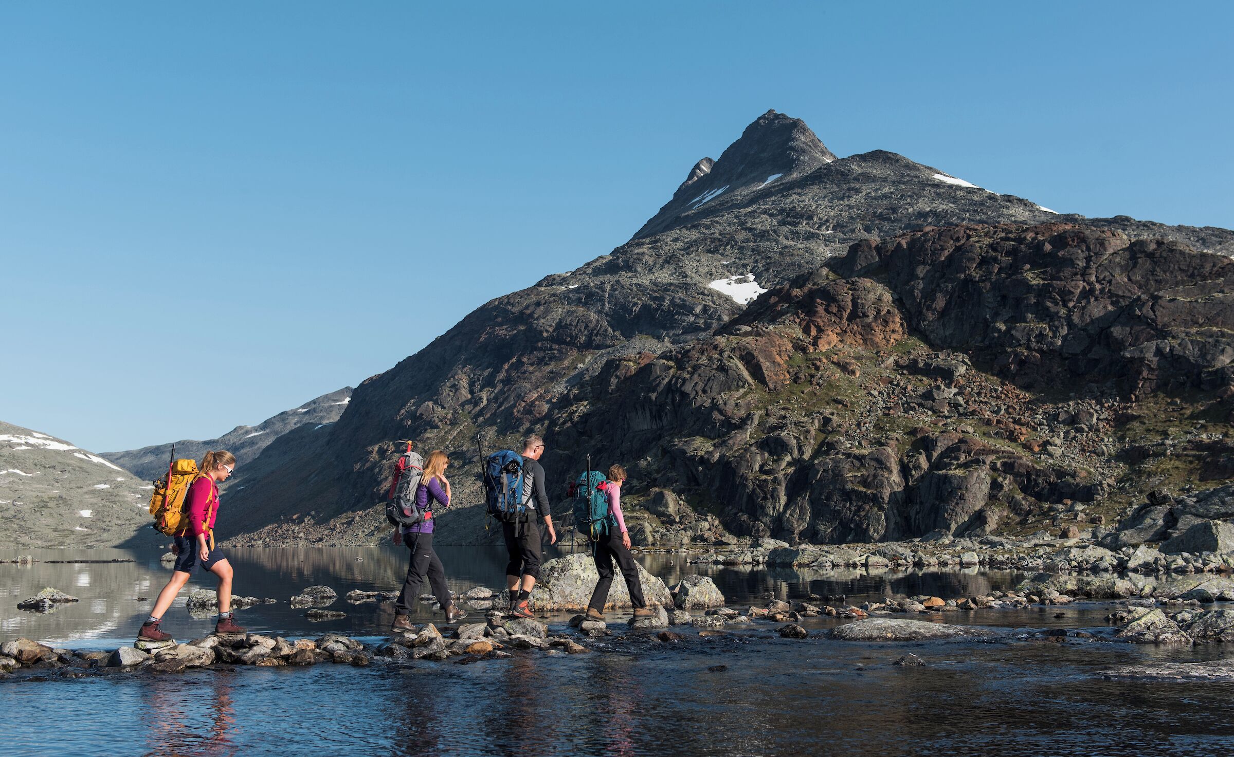 A group of hikers crossing a rocky stream with mountains in the background.