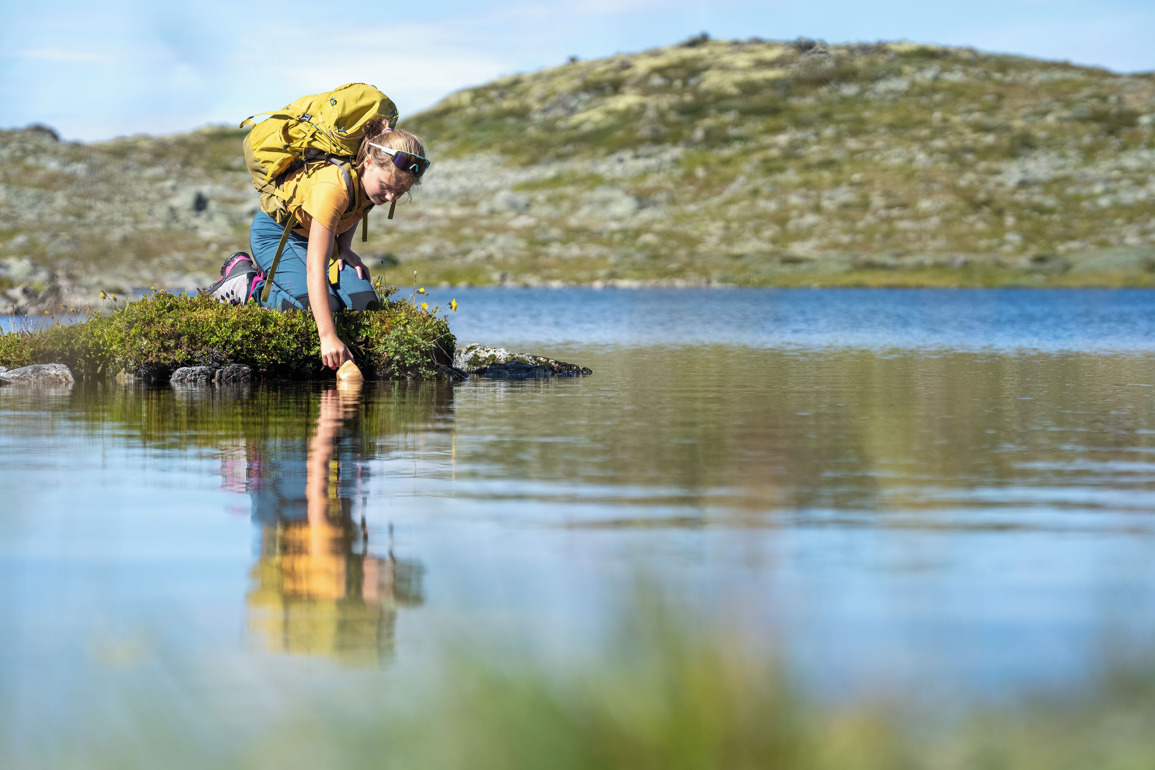 Une personne vêtue d’un t-shirt jaune et d’un short bleu, à genoux près d’un lac paisible, porte un sac à dos et est entourée de verdure et d’un paysage vallonné.