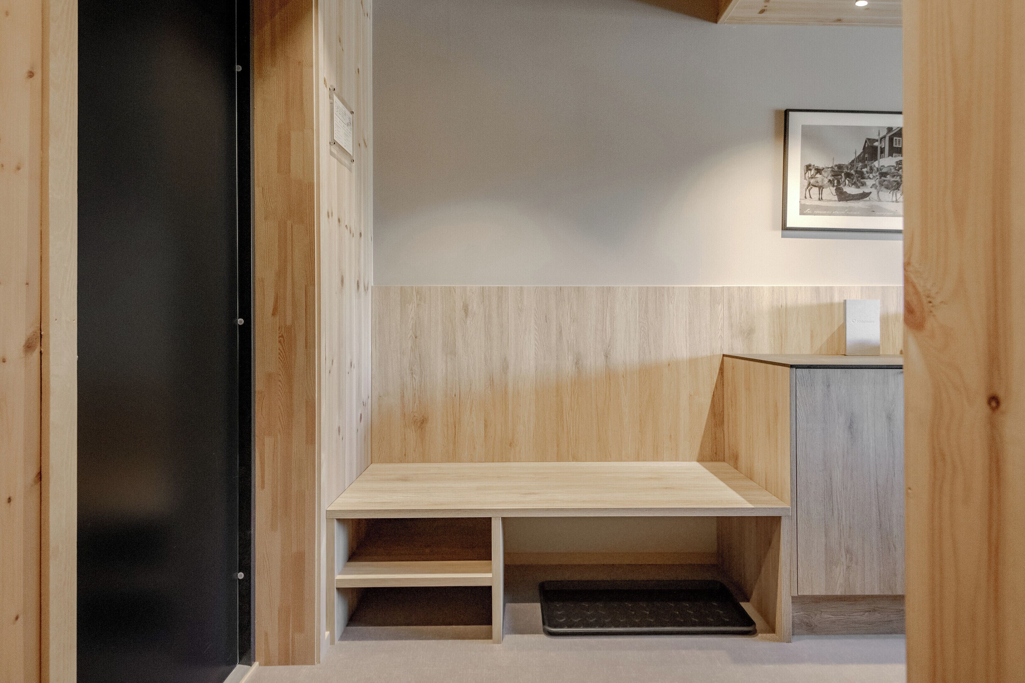 A wooden bench with a low backrest and a small shelf underneath, positioned against a wall with a light-colored panel.