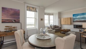 A living room with a round table, two chairs, a sofa, and a view of the beach through large windows. - A 4-bedroom luxury holiday home with enchanting sea views across Milsey Bay Beach (North Berwick)