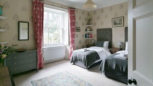 Un dormitorio con cama, cómoda, ventana con cortinas, radiador y mesita de noche con lámpara.