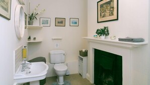 Un baño con un lavamanos blanco, inodoro y chimenea verde. Hay cuadros en la pared y una planta en maceta en el estante.