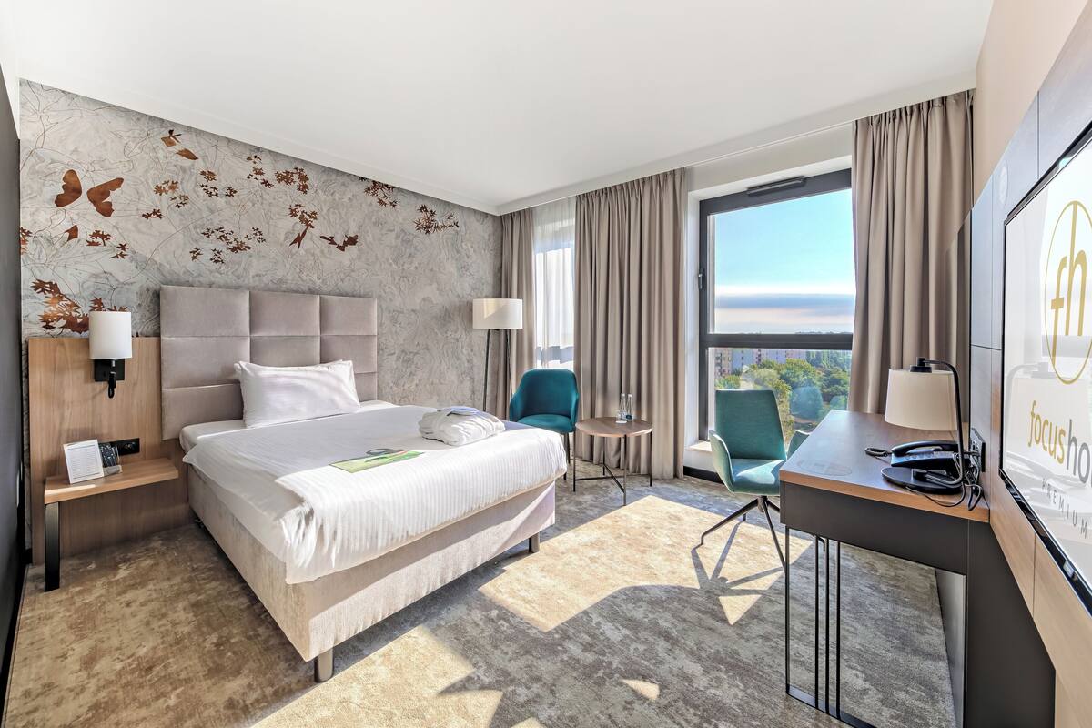 A hotel room with a bed, a desk, a chair, a TV, and a view of the city.