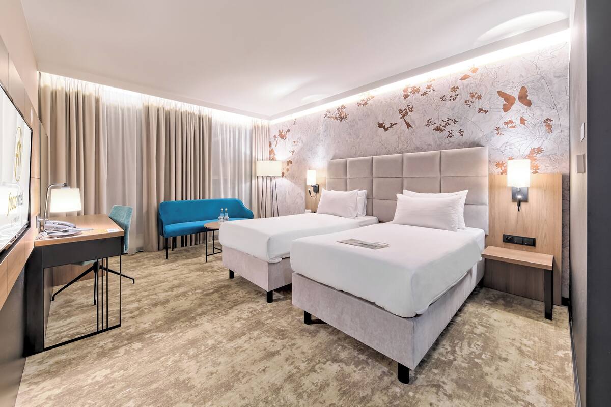 A hotel room with two beds, a desk, a blue chair, and a large window with curtains.