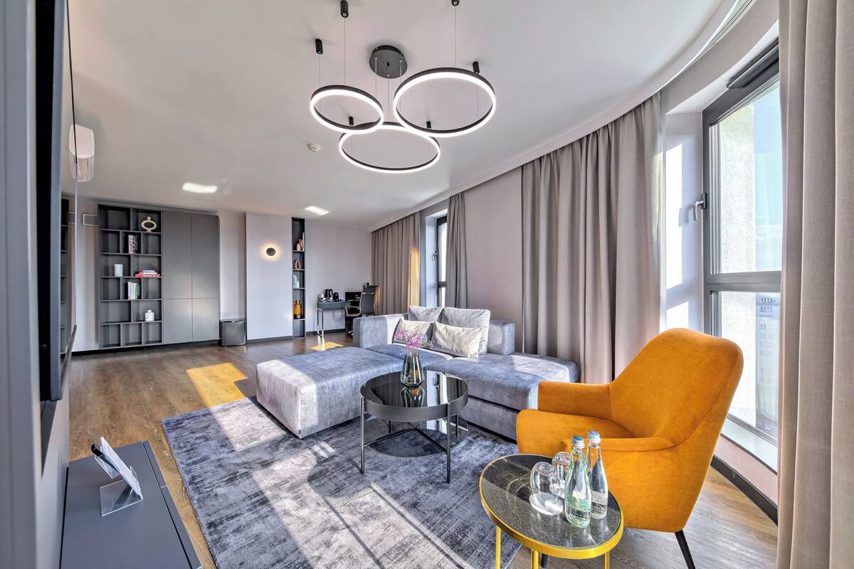 A modern living room with a grey sofa, a round coffee table, and an orange armchair. There is a large window with curtains, a built-in bookshelf, and a kitchen area in the background.