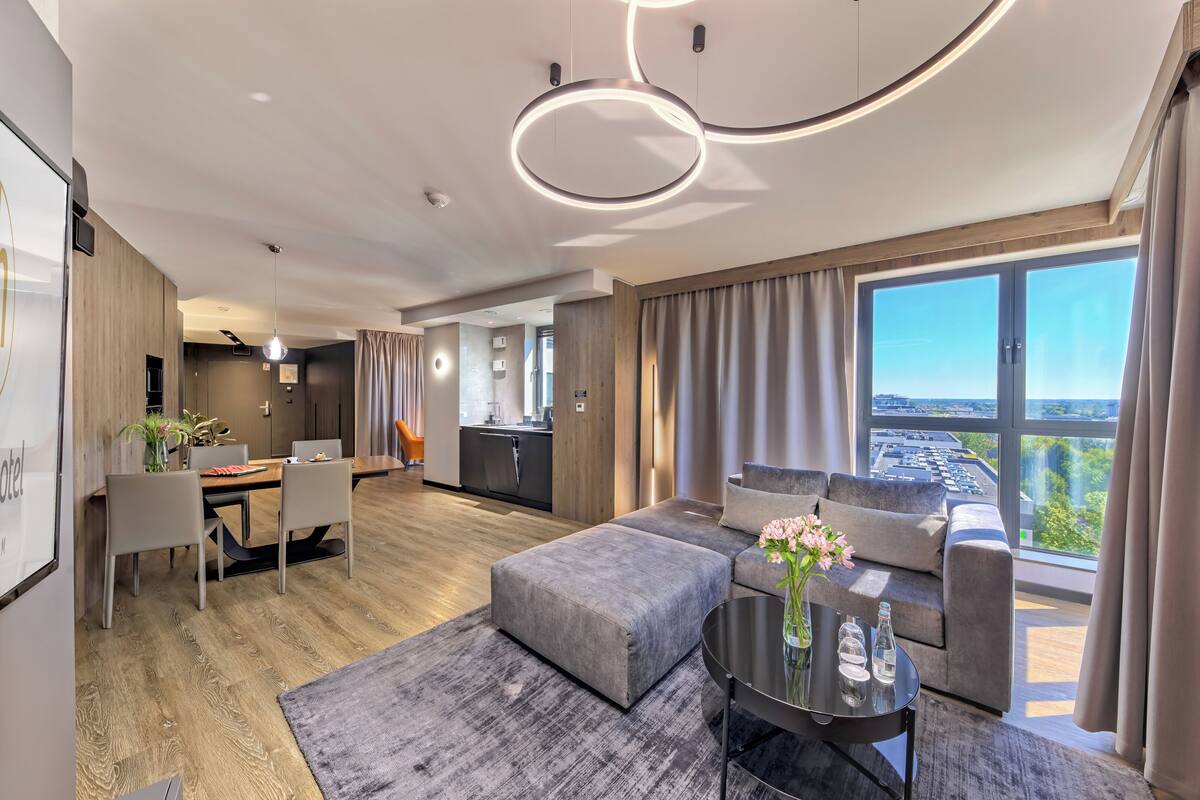 A modern hotel room with a dining area, a sofa, and a large window offering a city view.