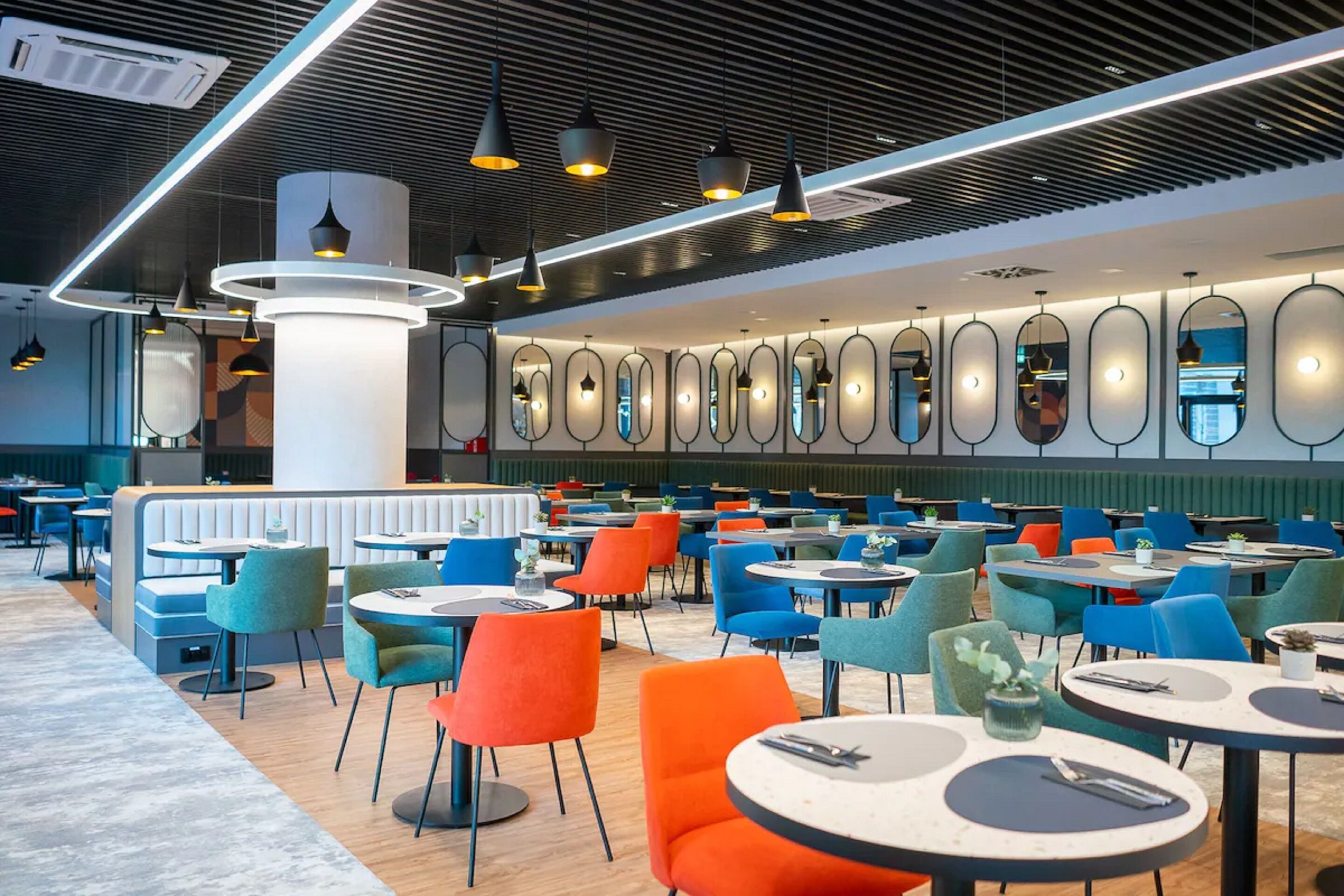 Een modern restaurant met ronde tafels en kleurrijke stoelen, een hoge plafond met verlichtingsarmaturen en een muur met ovale spiegels.