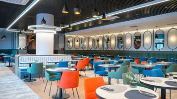 Een modern restaurant met ronde tafels en kleurrijke stoelen, een hoge plafond met verlichtingsarmaturen en een muur met ovale spiegels.