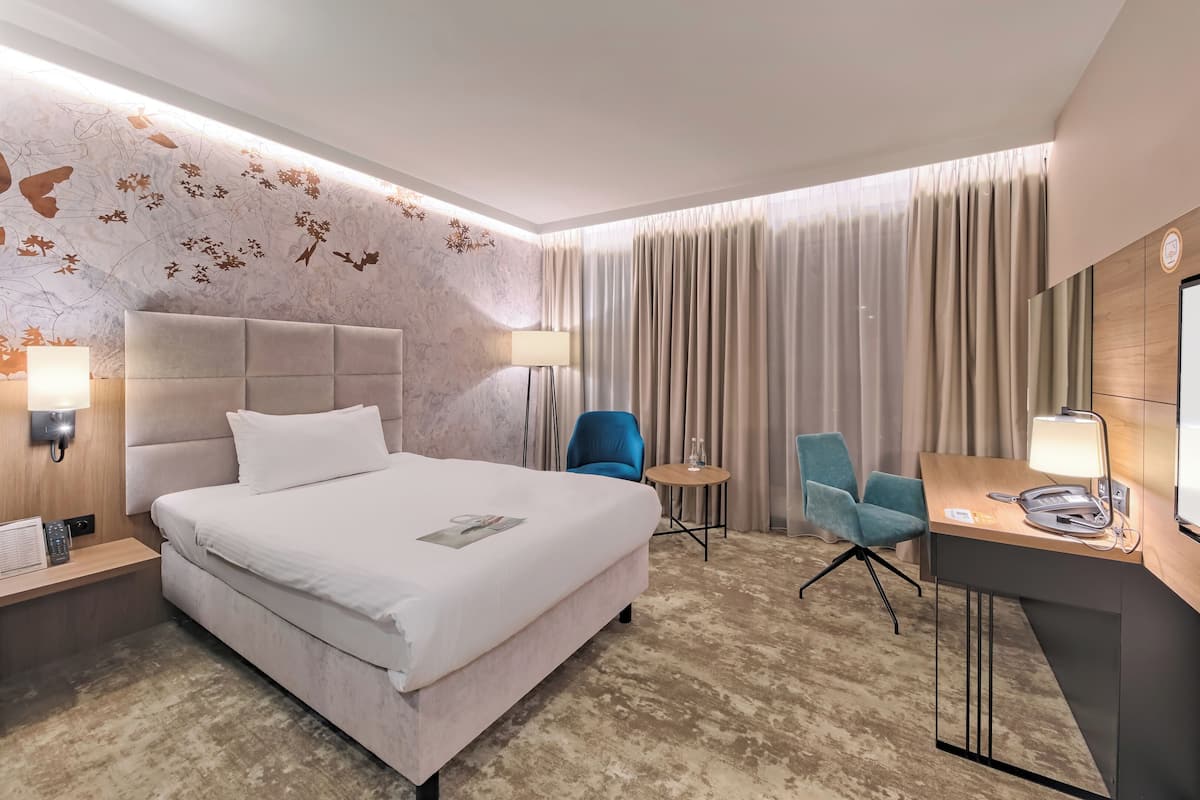 A hotel room with a large bed, a desk with a lamp, two chairs, and a wall-mounted TV.
