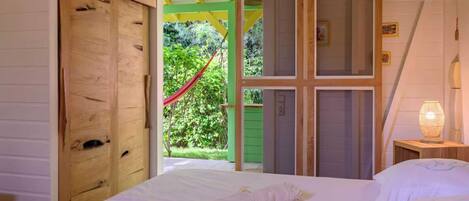 Quarto com cama, porta de madeira e vista para um jardim com rede.