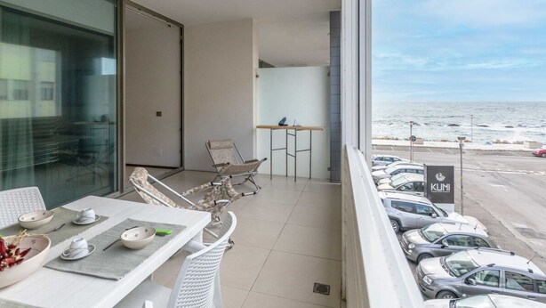Een balkon met een gedekte eettafel, een stoel en uitzicht op de oceaan en geparkeerde auto's.