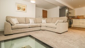 A modern living room with a beige sectional sofa, a glass coffee table, and a kitchen area with a washing machine. - 1 The Riverside (Barnard Castle)