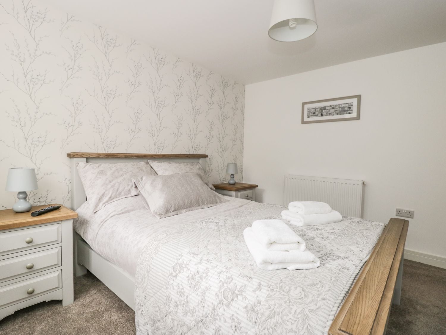 Ein ordentlich gemachtes Bett mit einem blumigem Überwurf und zwei Kissen, ein Nachttisch mit Lampe sowie ein gerahmtes Bild an der Wand.