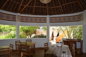 A dining area with a thatched roof, wooden furniture, and large windows offering views of greenery. - Zawadi Residence (Watamu)