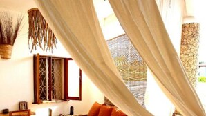Ein gemütliches Zimmer mit einem Bett, einem Holztisch und einem Stuhl. Ein Fenster mit Vorhängen und eine Pflanze hängen von der Decke.